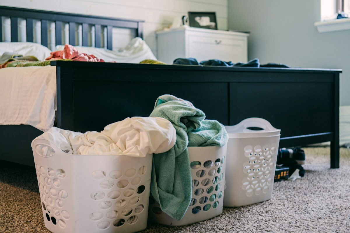 Three white laundry baskets of washing at the foot of a bed