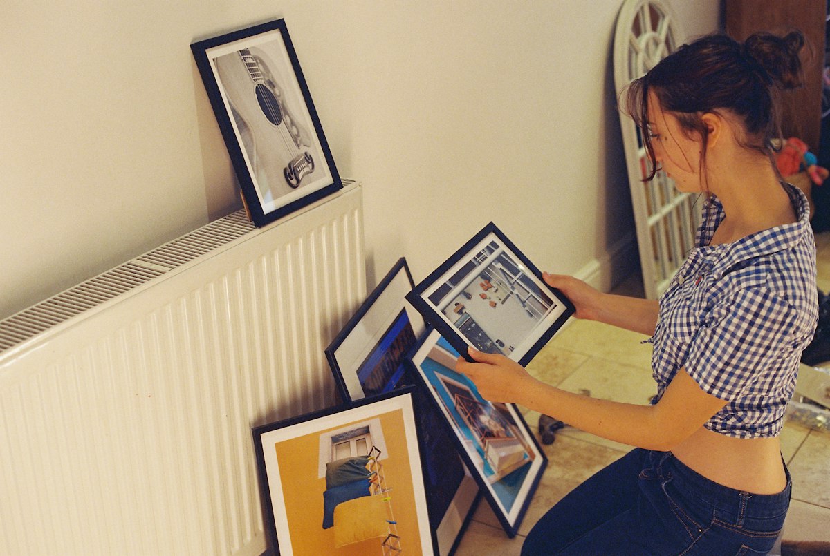A woman kneeling by a radiator sorting through framed pictures